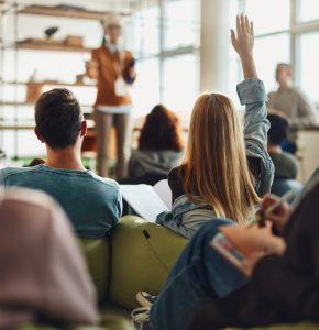 Back view of female student raising her hand to ask a question while having a class in a casual classroom.